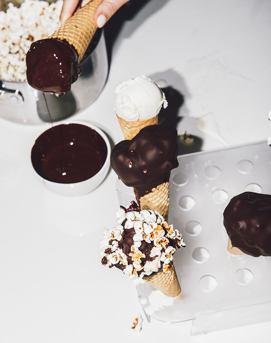 Ice cream cones with chocolate dip and popcorn topping on a white background, with a hand holding the cone.
