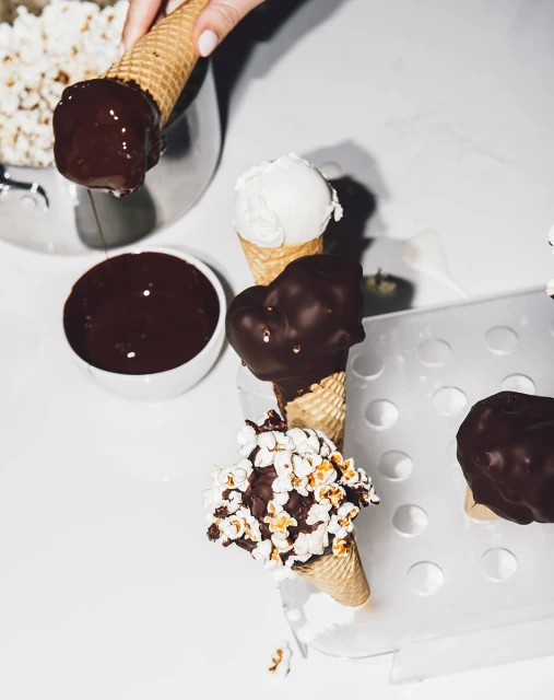 Ice cream cones with chocolate dip and popcorn topping on a white background, with a hand holding the cone.