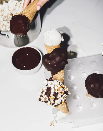 Ice cream cones with chocolate dip and popcorn topping on a white background, with a hand holding the cone.