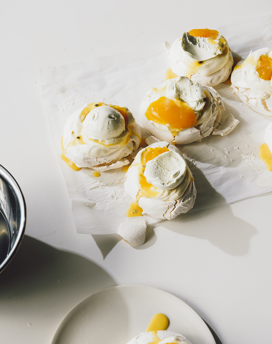 Overhead shot of meringue desserts with white topping and yellow sauce on parchment paper and a white surface with a metal bowl.