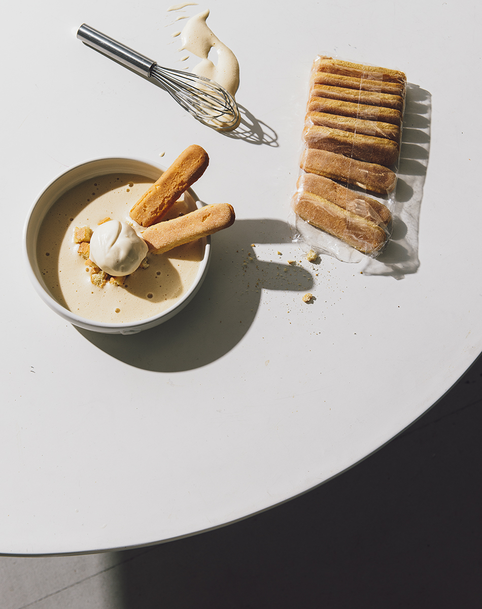 Overhead shot of tiramisu ingredients on a white table: ladyfingers, cream, dessert sauce, and whisk casting a shadow.