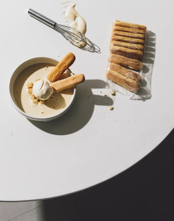 Overhead shot of tiramisu ingredients on a white table: ladyfingers, cream, dessert sauce, and whisk casting a shadow.