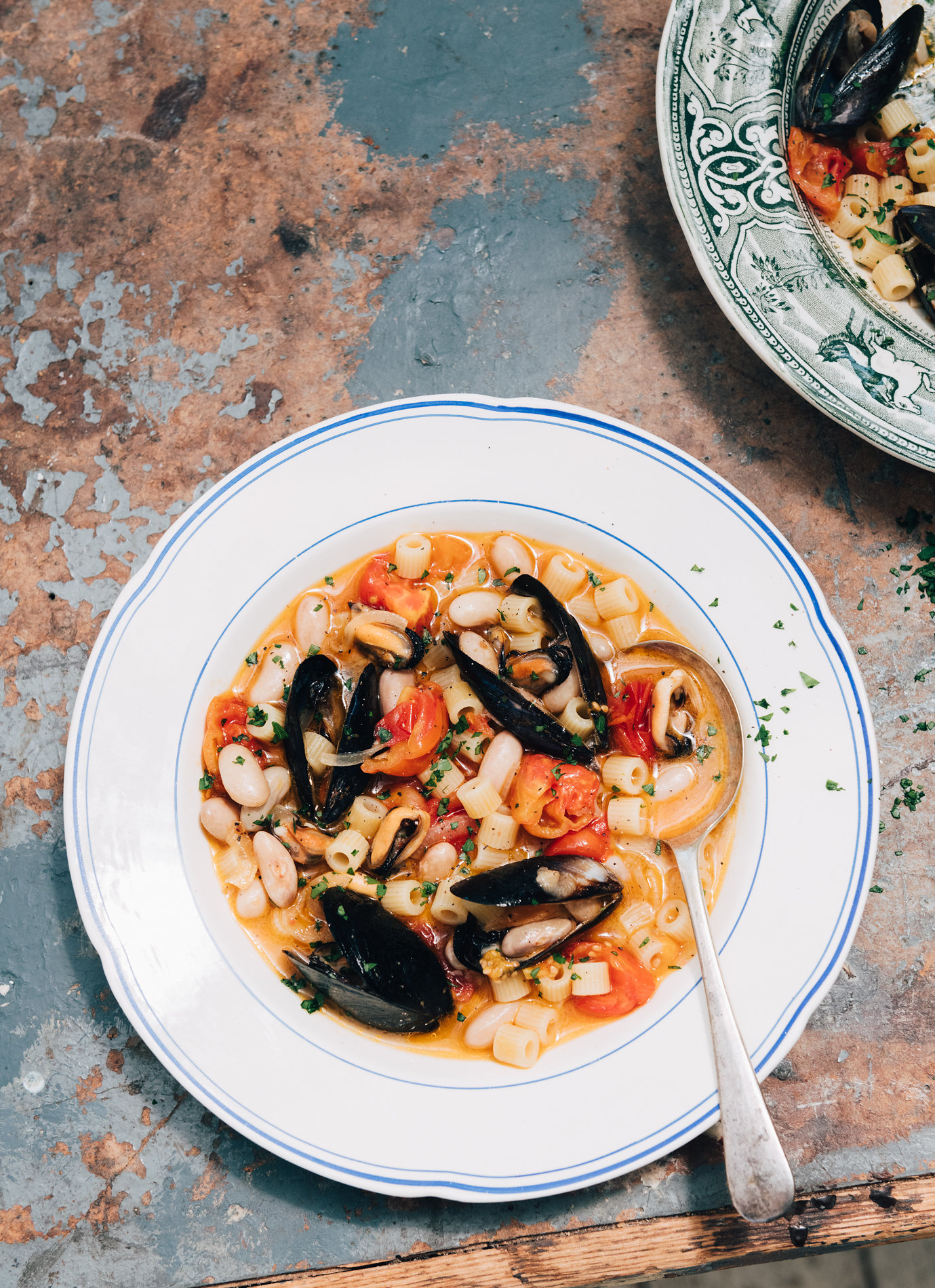 Overhead shot of mussel and pasta soup with tomatoes and beans in white bowl with spoon, set on rustic textured surface.