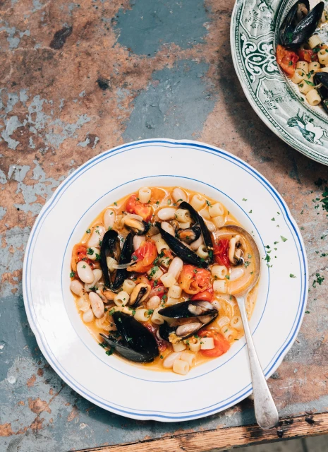 Overhead shot of mussel and pasta soup with tomatoes and beans in white bowl with spoon, set on rustic textured surface.