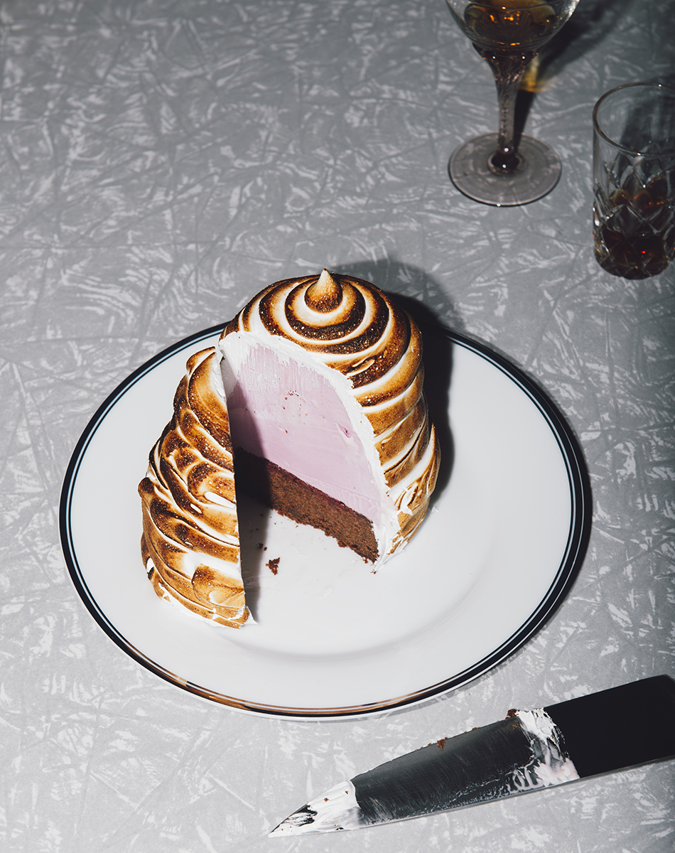 Elevated view of a sliced Baked Alaska dessert with toasted meringue, pink ice cream, chocolate cake, and serving knife on a tablecloth.