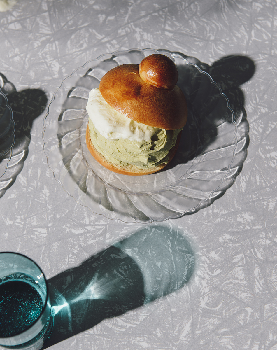 Brioche-style pastry with cream and green filling on a clear glass plate. A textured tablecloth and glass add to the table setting.