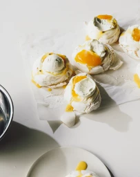 Several small meringue desserts topped with cream and passionfruit curd arranged on parchment paper