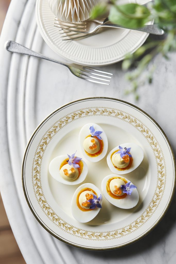Overhead shot of deviled eggs with caviar and purple flowers on a decorative plate set on a marble table with silverware.