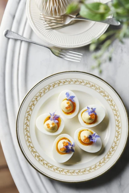 Overhead shot of deviled eggs with caviar and purple flowers on a decorative plate set on a marble table with silverware.