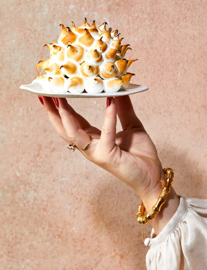 A hand holds a small white plate topped with a torched meringue dessert, set against a textured pink backdrop. Gold bracelet visible.