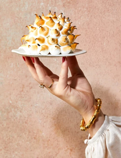 A hand holds a small white plate topped with a torched meringue dessert, set against a textured pink backdrop. Gold bracelet visible.