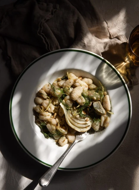 Overhead shot of white beans, fennel, and herbs served on a white plate with green trim, accompanied by a silver fork.