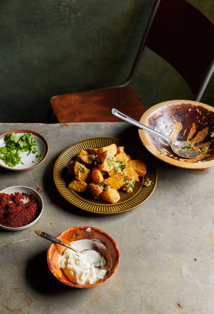 Overhead view of a rustic table with bowls of spiced potatoes, white sauce, herbs, and a metal spoon, arranged for a culinary photoshoot.