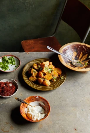 Overhead view of a rustic table with bowls of spiced potatoes, white sauce, herbs, and a metal spoon, arranged for a culinary photoshoot.