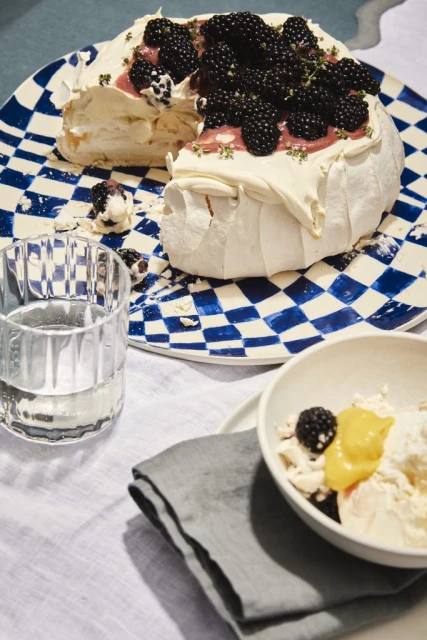 Overhead shot of a pavlova cake with blackberries, served on a checkered plate with a glass of water and linen napkin.