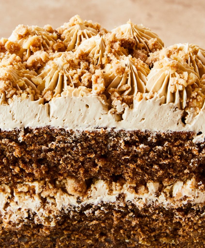 Close-up of a slice of frosted chocolate cake featuring layers of cake and cream, topped with crumbled cake pieces and light-brown frosting.