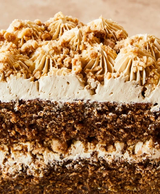 Close-up of a slice of frosted chocolate cake featuring layers of cake and cream, topped with crumbled cake pieces and light-brown frosting.