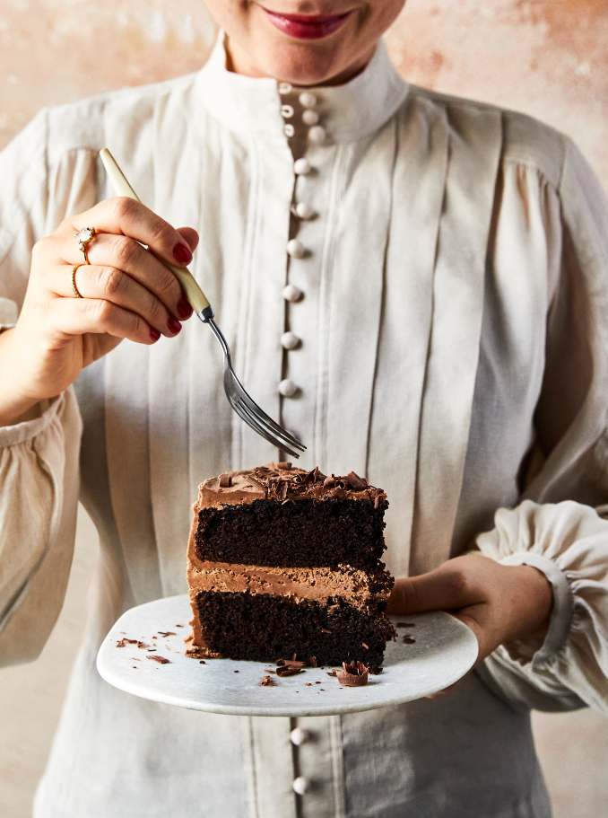 Woman holds slice of chocolate cake on a plate, ready to eat with a fork, in a brightly lit, close-up food photography studio shot.