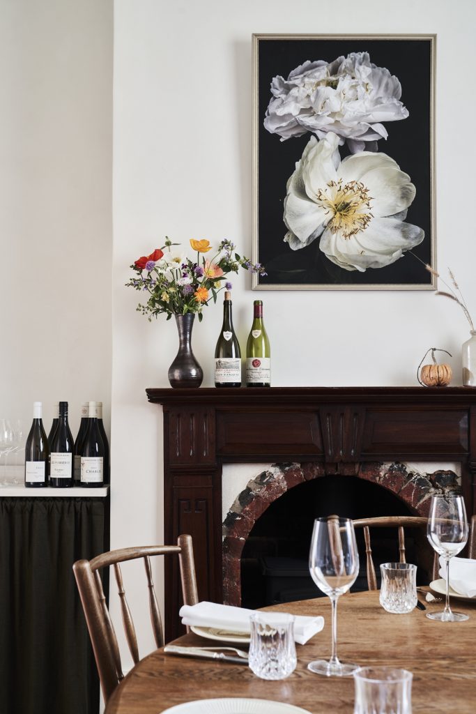 Dining room interior with a wooden table set with wine glasses and plates, flowers, and a floral art print above a fireplace.