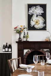 Interior dining space with a dark wood mantel, wine bottles, fresh wildflowers, and a large white floral art print on the wall.