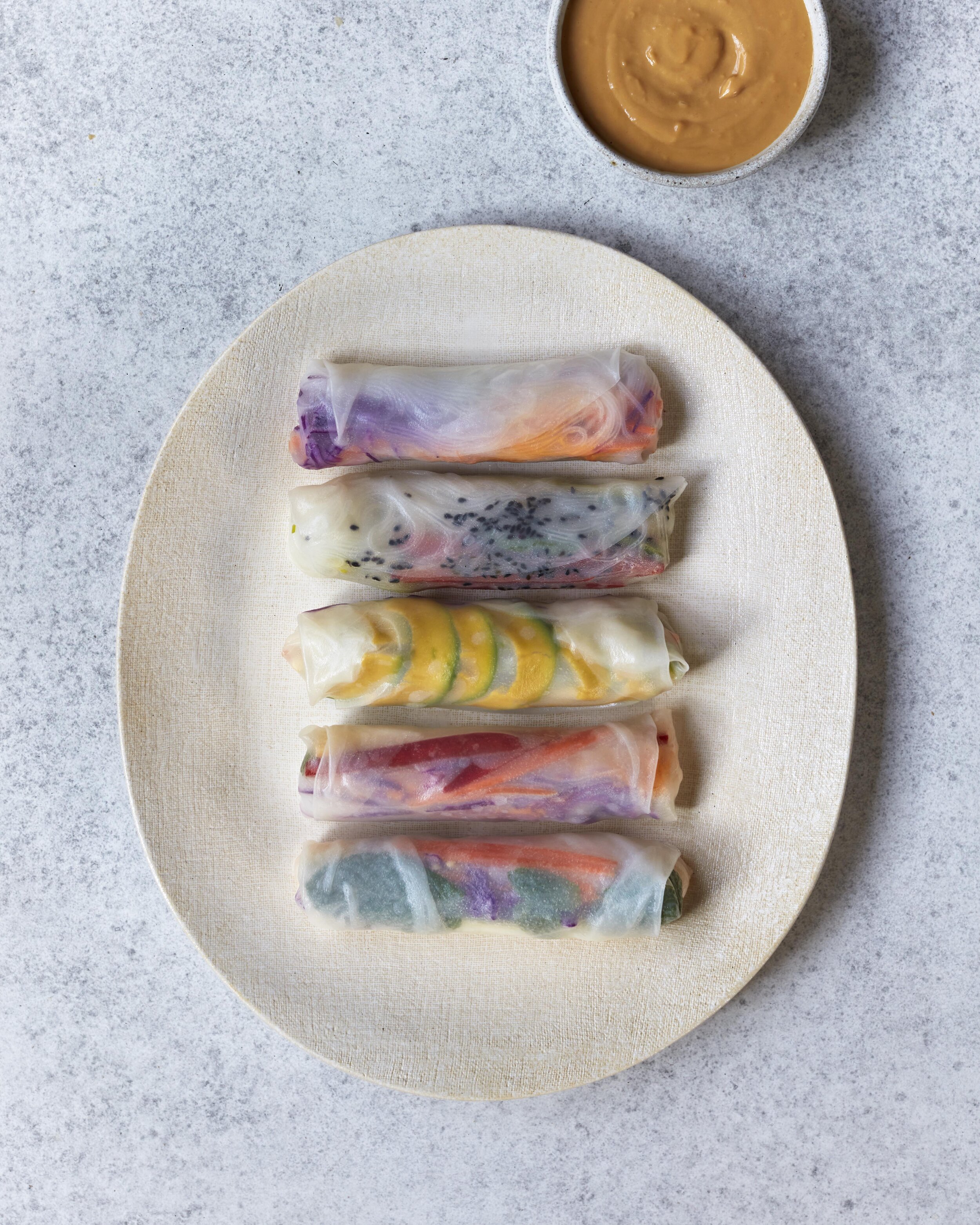 Overhead shot of spring rolls with colorful fillings arranged on a plate, accompanied by a bowl of peanut sauce, against a neutral backdrop.