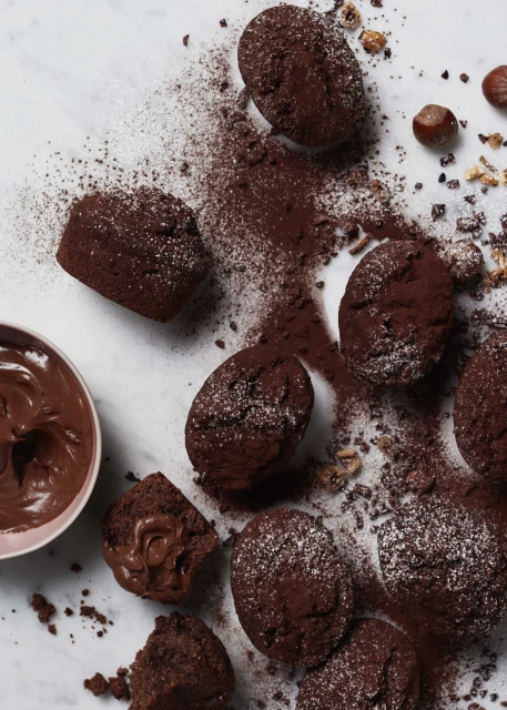 Overhead shot of chocolate muffins with cocoa powder dusting, chocolate sauce and hazelnuts on white marble surface, food photography.