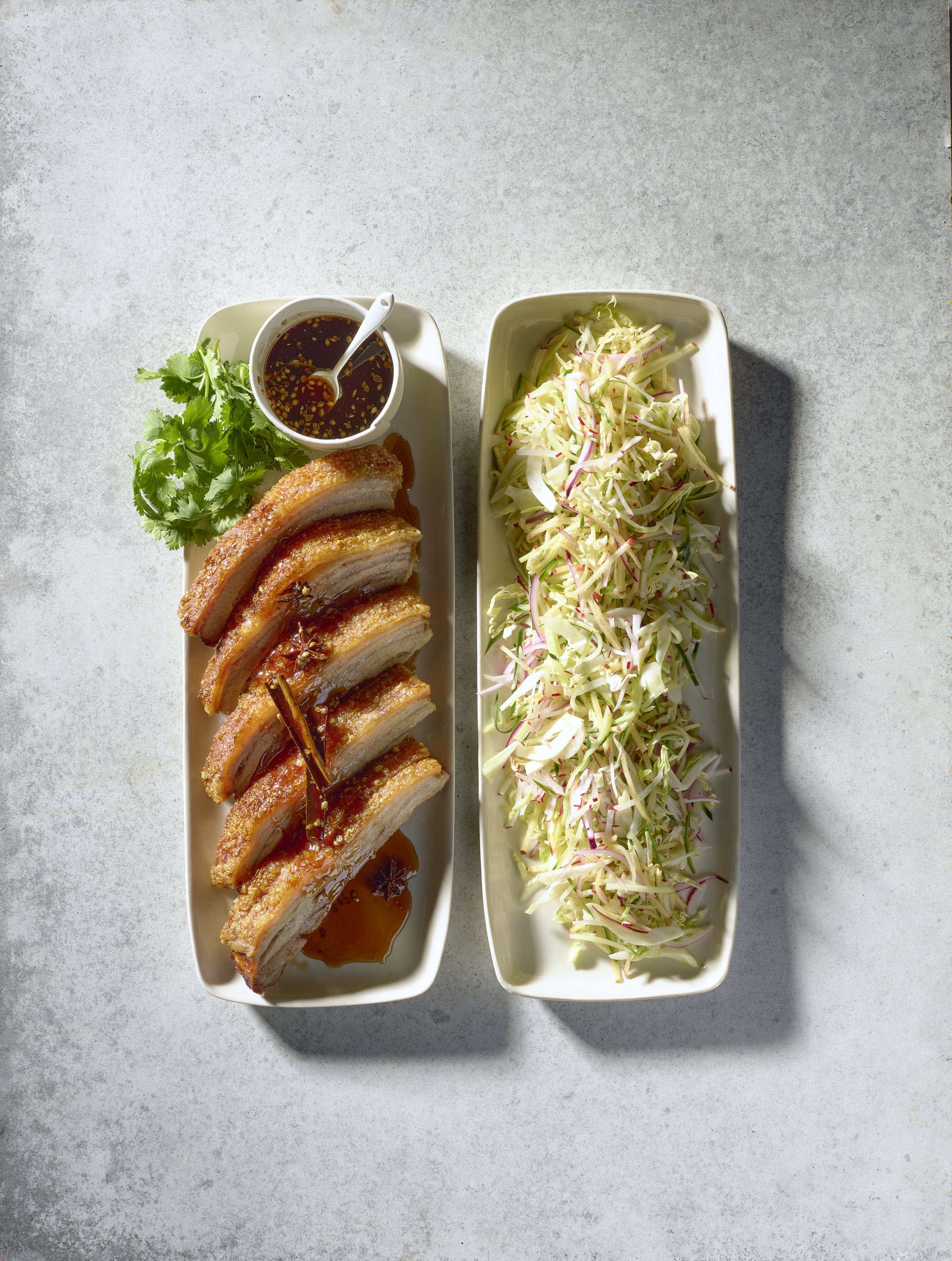 Overhead shot of crispy pork belly with dipping sauce and coleslaw on rectangular white plates set on a mottled grey surface.