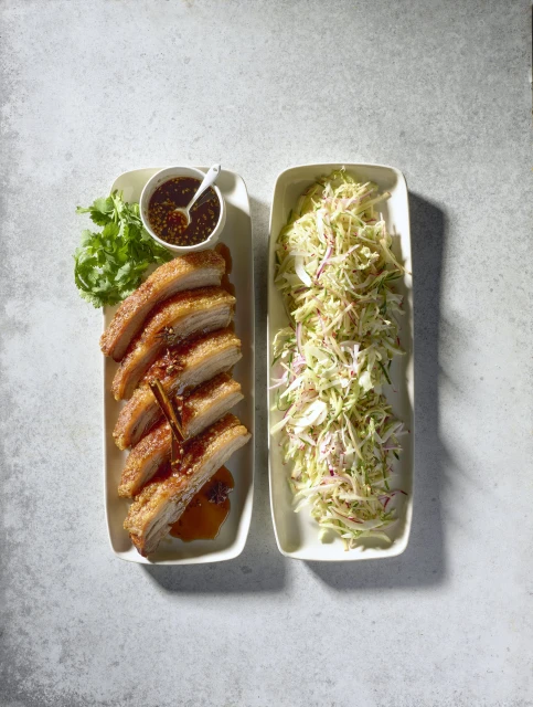 Overhead shot of crispy pork belly with dipping sauce and coleslaw on rectangular white plates set on a mottled grey surface.