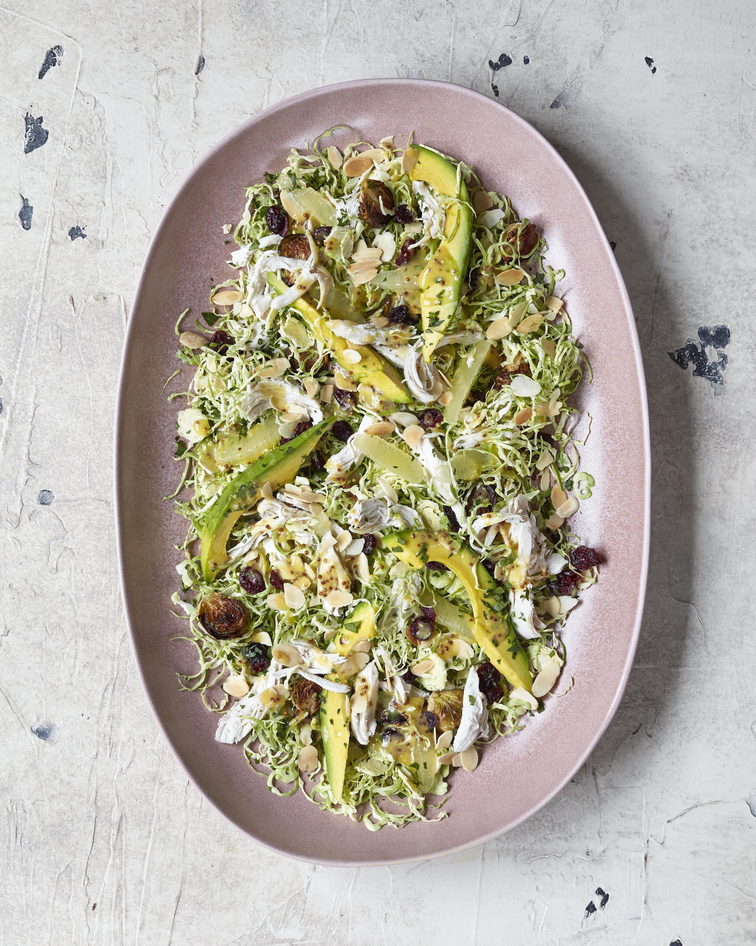 Overhead shot of shredded brussel sprouts and avocado salad on a pink oval platter, accented with toasted almonds and dried cranberries.