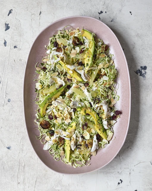 Overhead shot of shredded brussel sprouts and avocado salad on a pink oval platter, accented with toasted almonds and dried cranberries.