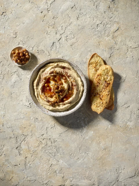 Overhead shot of hummus topped with chickpeas in a white bowl, accompanied by a side of chickpeas and pieces of pita bread.