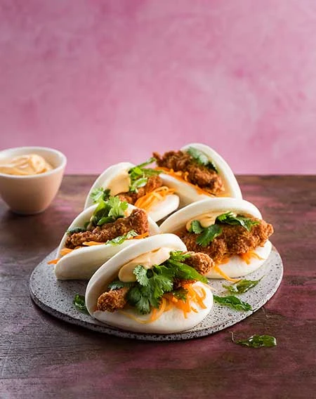 Several Taiwanese bao buns filled with fried chicken, fresh cilantro, and shredded carrots are arranged on a gray serving plate.