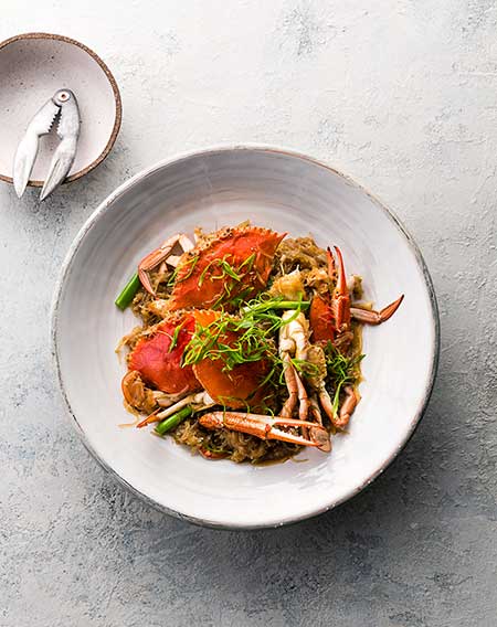 Overhead shot of cooked crab dish on a white plate, garnished with herbs, accompanied by crab cracker on mottled gray backdrop.