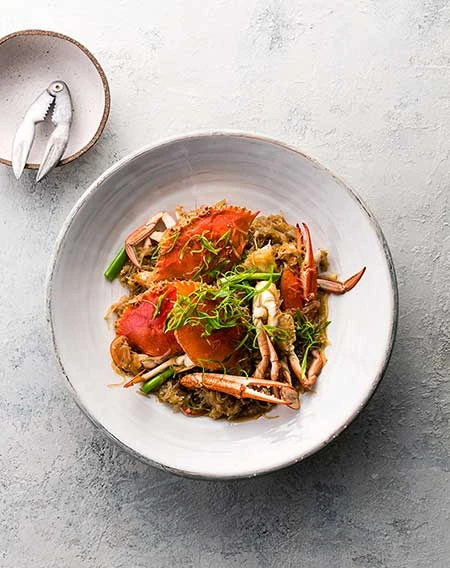 Overhead shot of cooked crab dish on a white plate, garnished with herbs, accompanied by crab cracker on mottled gray backdrop.
