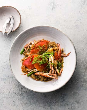Overhead shot of cooked crab dish on a white plate, garnished with herbs, accompanied by crab cracker on mottled gray backdrop.