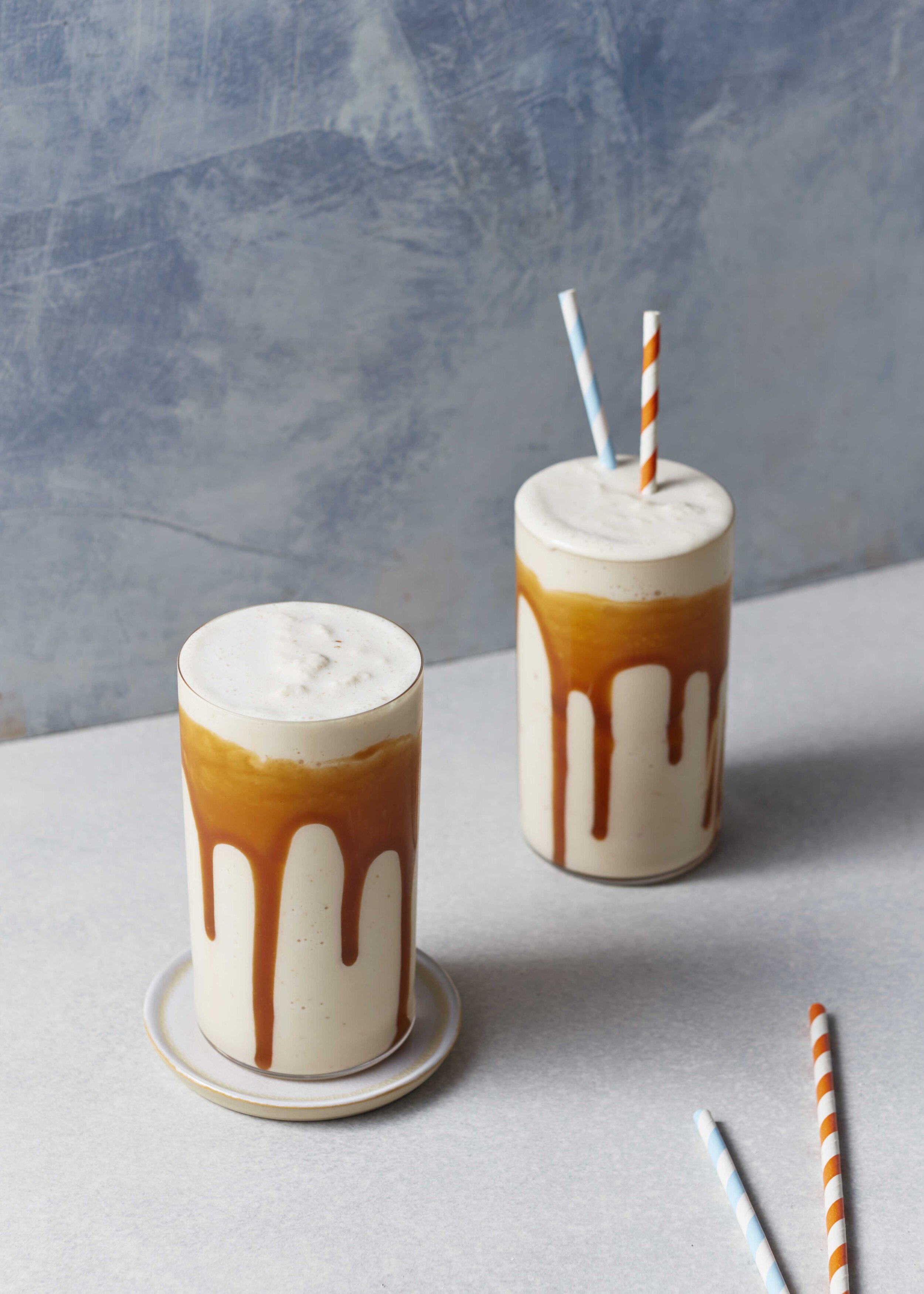 Two glasses of milkshake with caramel drizzle and striped straws on a light surface in front of a textured blue background.