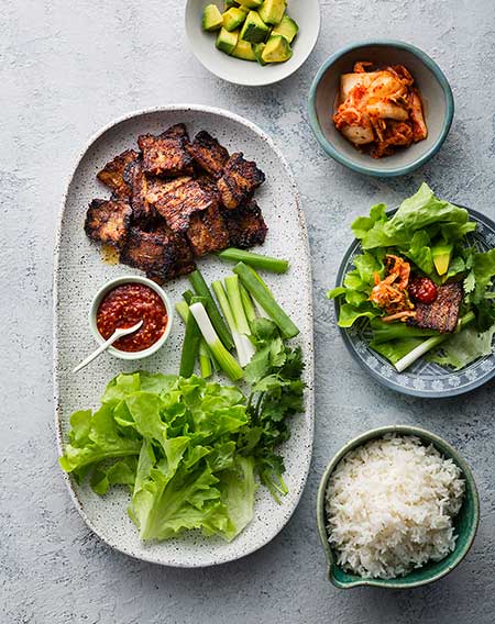 Overhead shot of Korean lettuce wraps with cooked pork belly, fresh lettuce, green onions, kimchi, avocado and spicy sauce on a grey surface.
