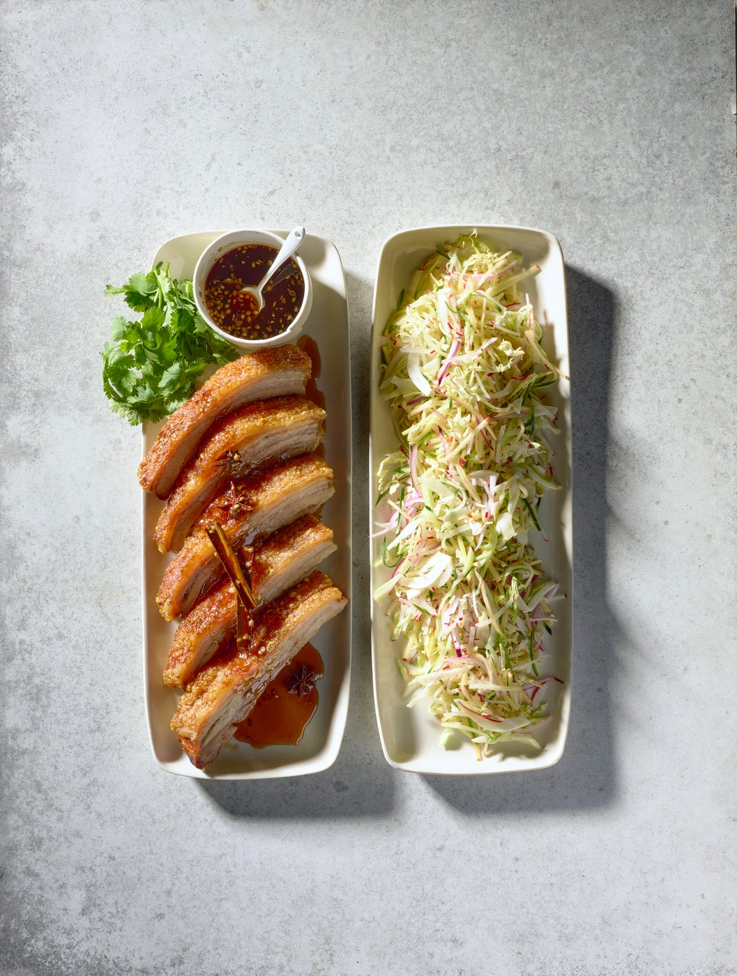 Two rectangular white dishes of sliced crispy pork belly with chili sauce and coleslaw styled on concrete
