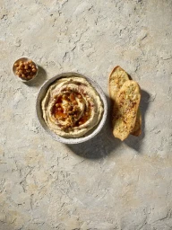 Bowl of creamy hummus topped with oil, spices, and chickpeas next to flatbread on textured stone