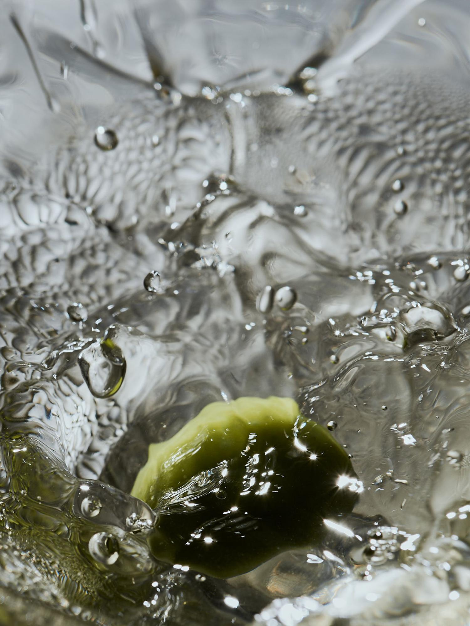 Close-up of a green dumpling or piece of food submerged in clear water, with water droplets and ripples visible.