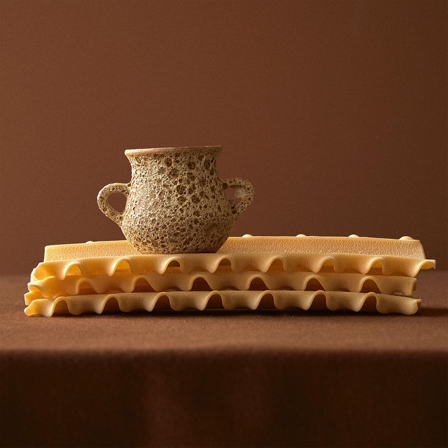 Close-up of uncooked lasagna noodles stacked with textured tan ceramic vase in still life arrangement against brown background.
