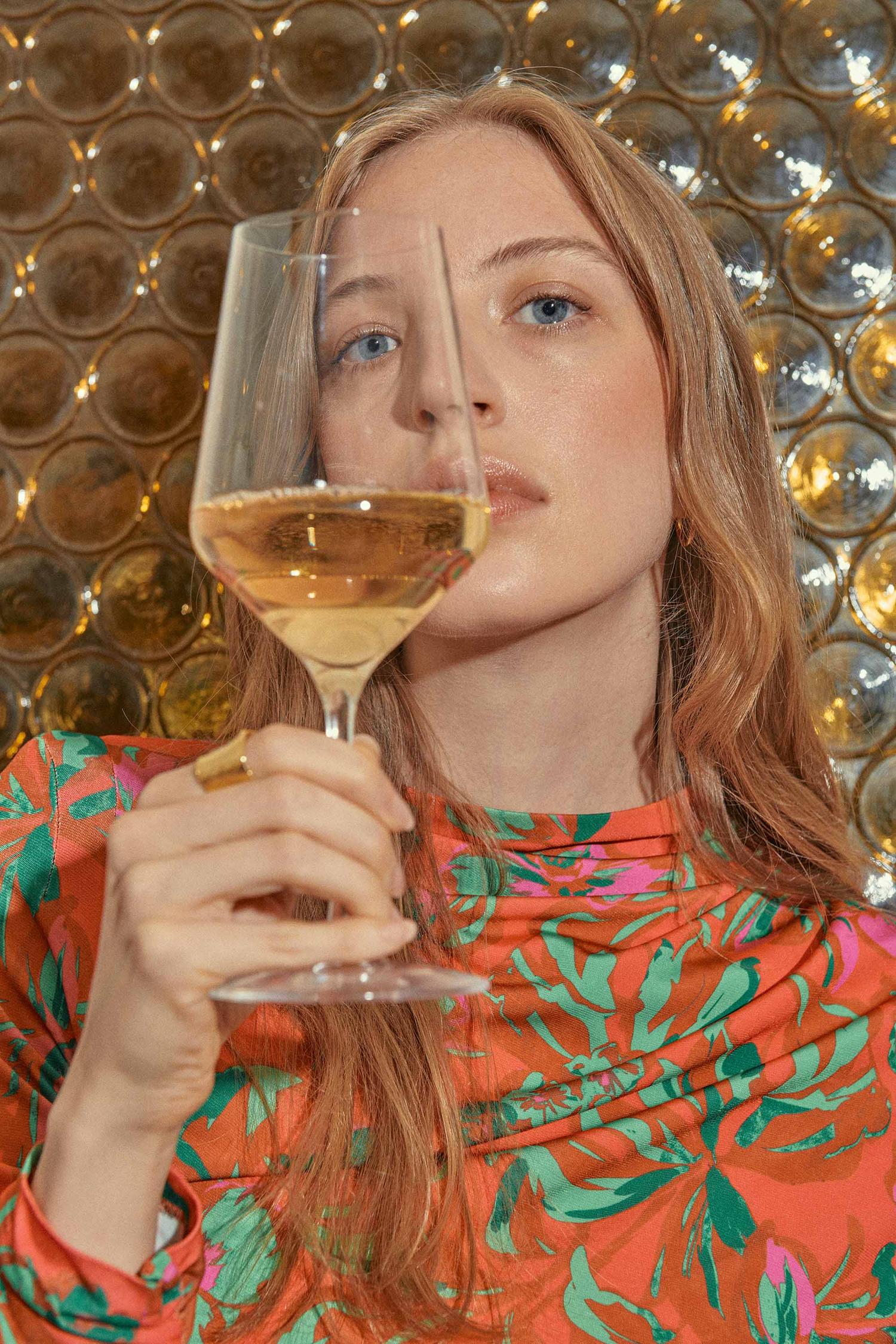 Woman holds glass of white wine in a floral-patterned top, set against a textured gold background. The frame is a close-up.