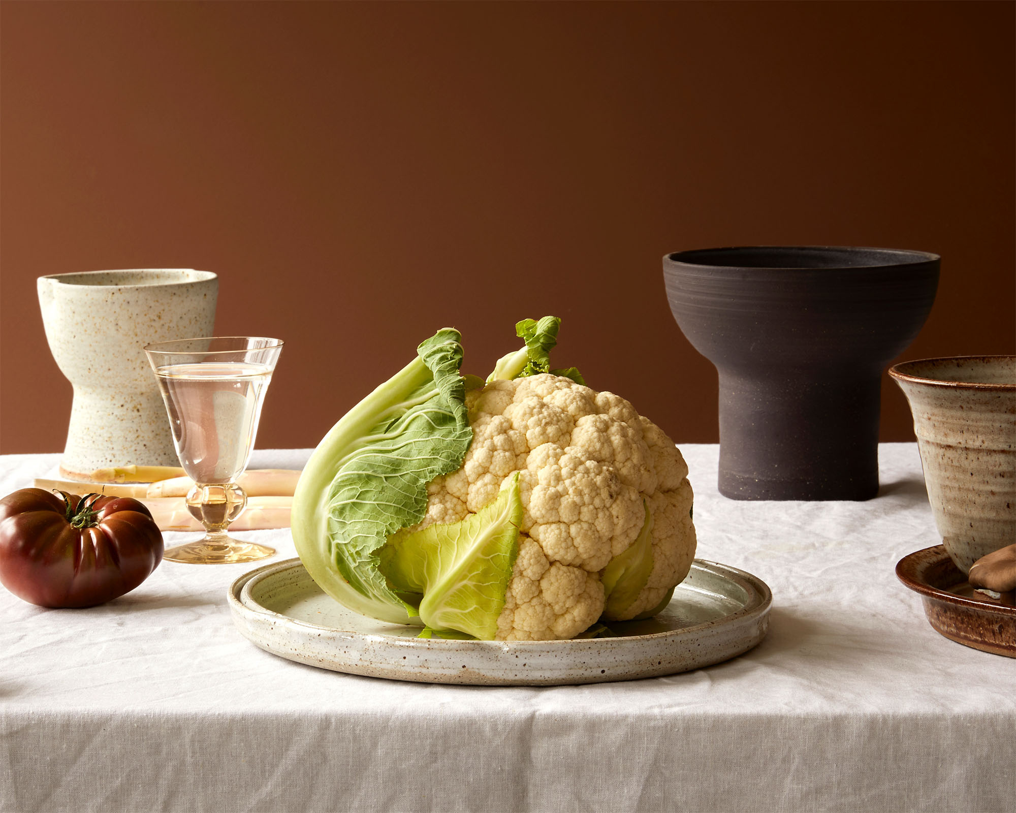 Still life photograph featuring a cauliflower centerpiece, dark tomato, drinking glass with water, and ceramic dishware on a white tablecloth.
