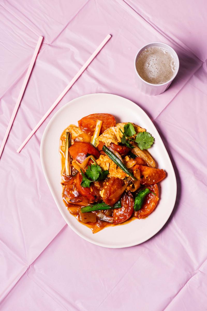 Overhead shot of a plated dish of tomato stir-fry with tofu and scallions, presented on a pink background with drinking straws and a beverage.