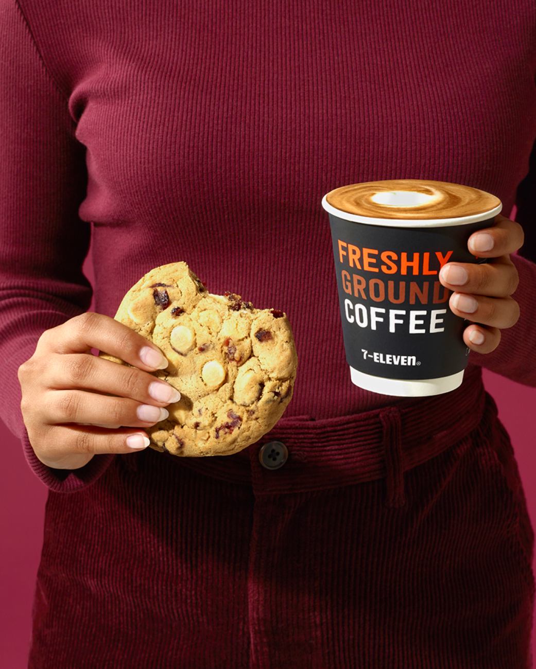 A person holds a large cookie with white chocolate chips and a 7-Eleven coffee cup against a reddish-purple background indoors.