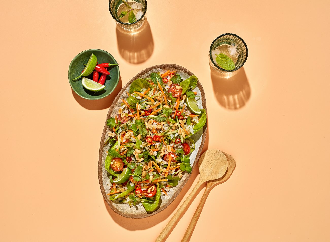 Overhead shot of a vibrant salad with various vegetables and nuts, served on a platter, alongside fresh limes, chilies, and drinks.