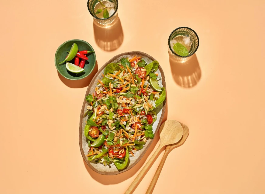 Overhead shot of a vibrant salad with various vegetables and nuts, served on a platter, alongside fresh limes, chilies, and drinks.