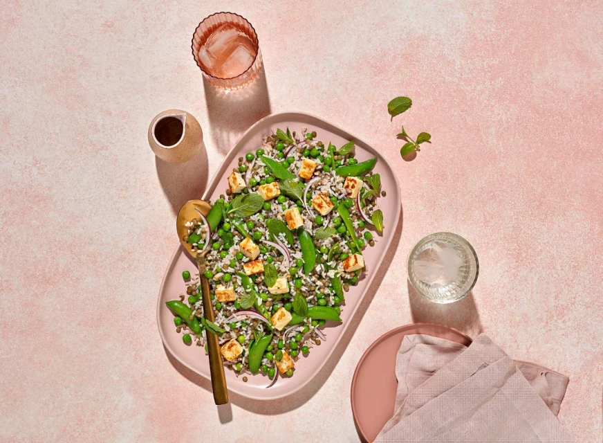 Overhead shot of a platter filled with pea and cheese salad, accompanied by drinks, a gold spoon, and a pale pink background.