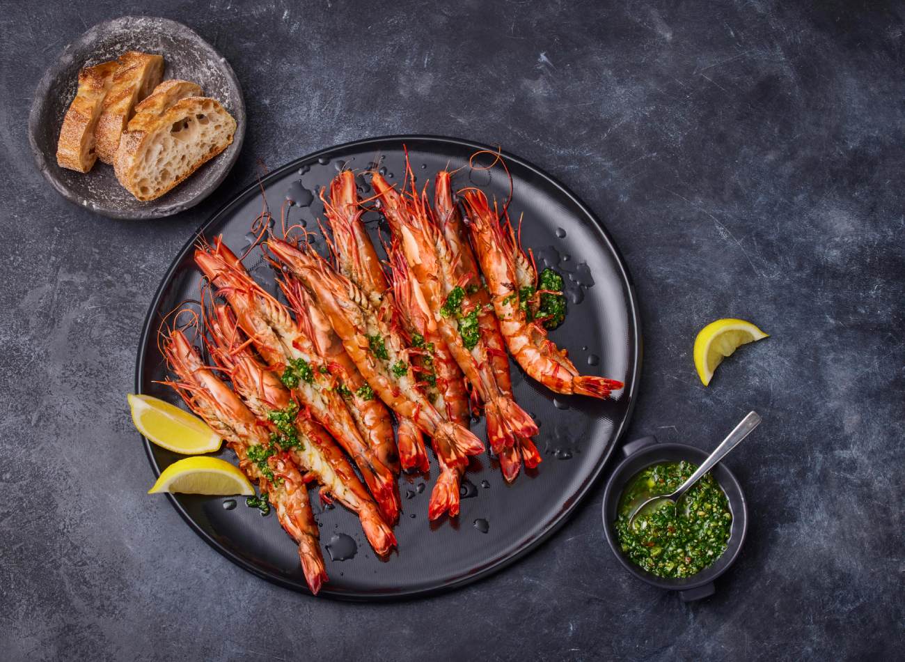 Overhead shot of grilled tiger prawns on a black plate, garnished with herbs and lemon wedges, alongside bread and dipping sauce.