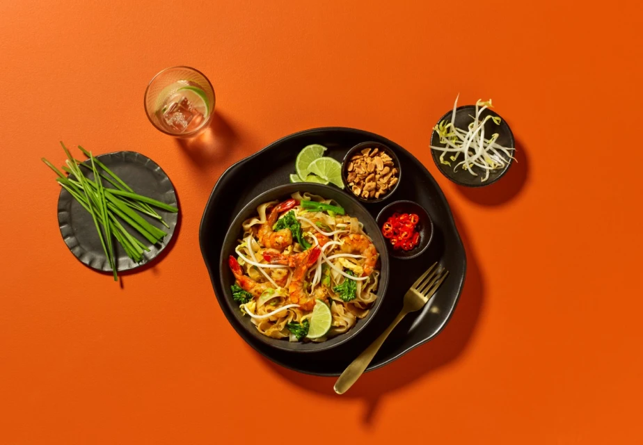 Overhead shot of shrimp pad thai in a black bowl, garnished with lime wedges, peanuts, sprouts, chili flakes, chives, and a cocktail.